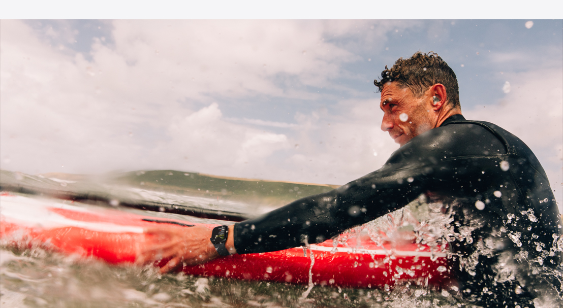 Um homem no mar usando um Apple Watch Series 10 com roupa de mergulho e empurrando uma prancha de surfe.