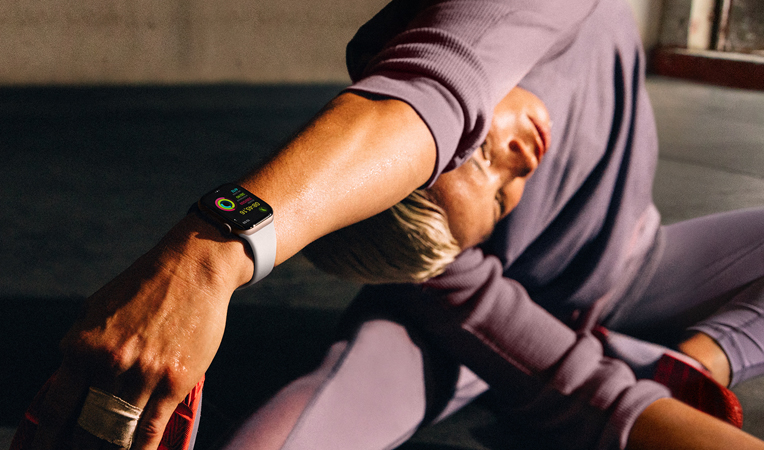 Une femme assise sur le sol étire son bras gauche au-dessus de sa tête vers sa jambe droite; elle porte une Apple Watch Series 10, l’écran affiche Exercice, Retour au calme, Activité, Anneaux d’activité