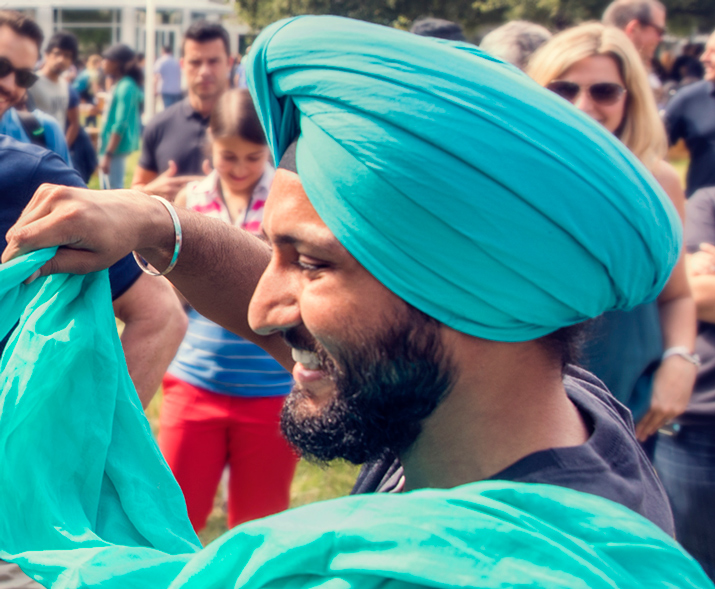 Photo of a man smiling, wearing a turban, wrapping another person’s turban.