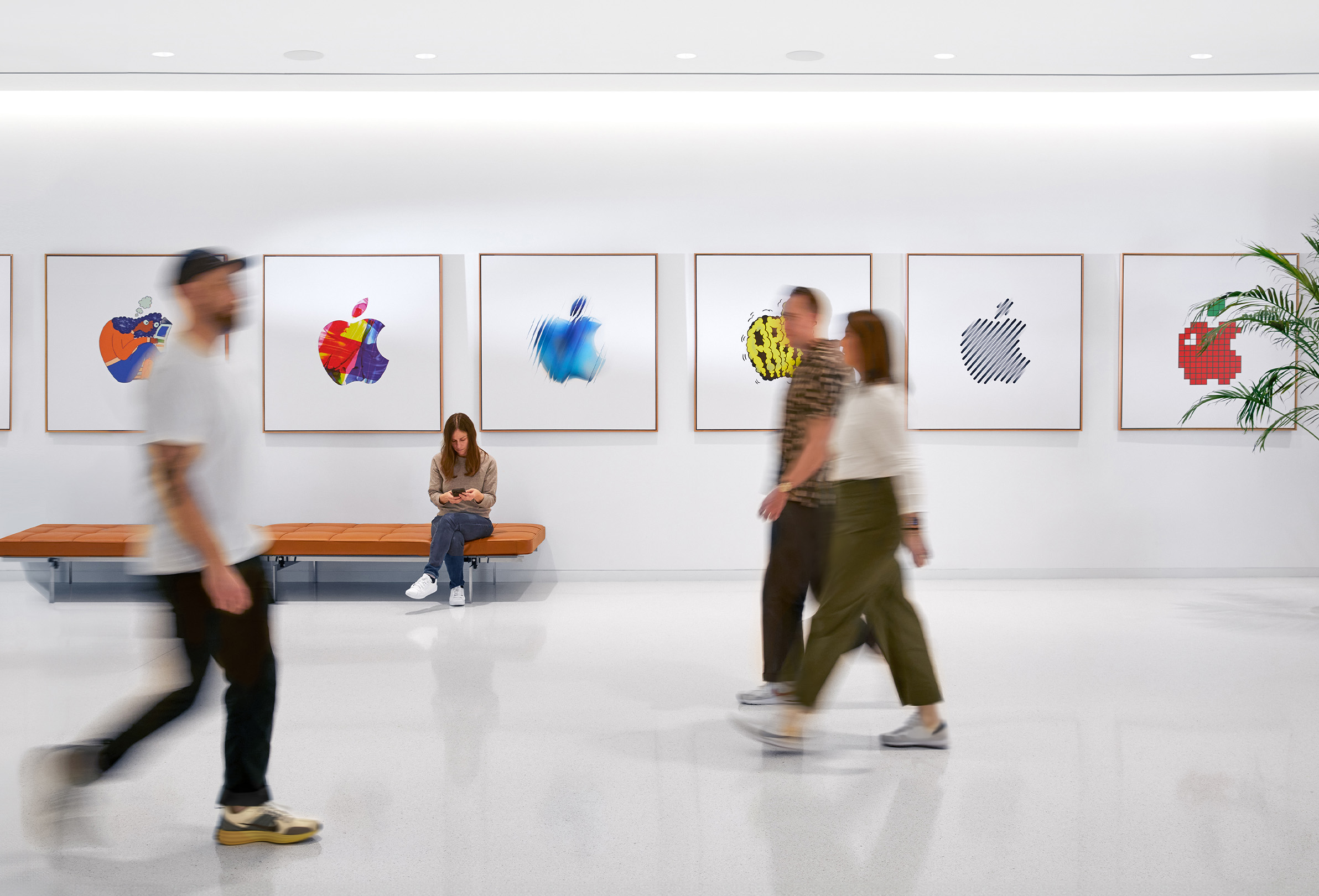 Three Apple colleagues walking past an interior wall with colorful Apple logos, and a fourth employee sitting on a bench.