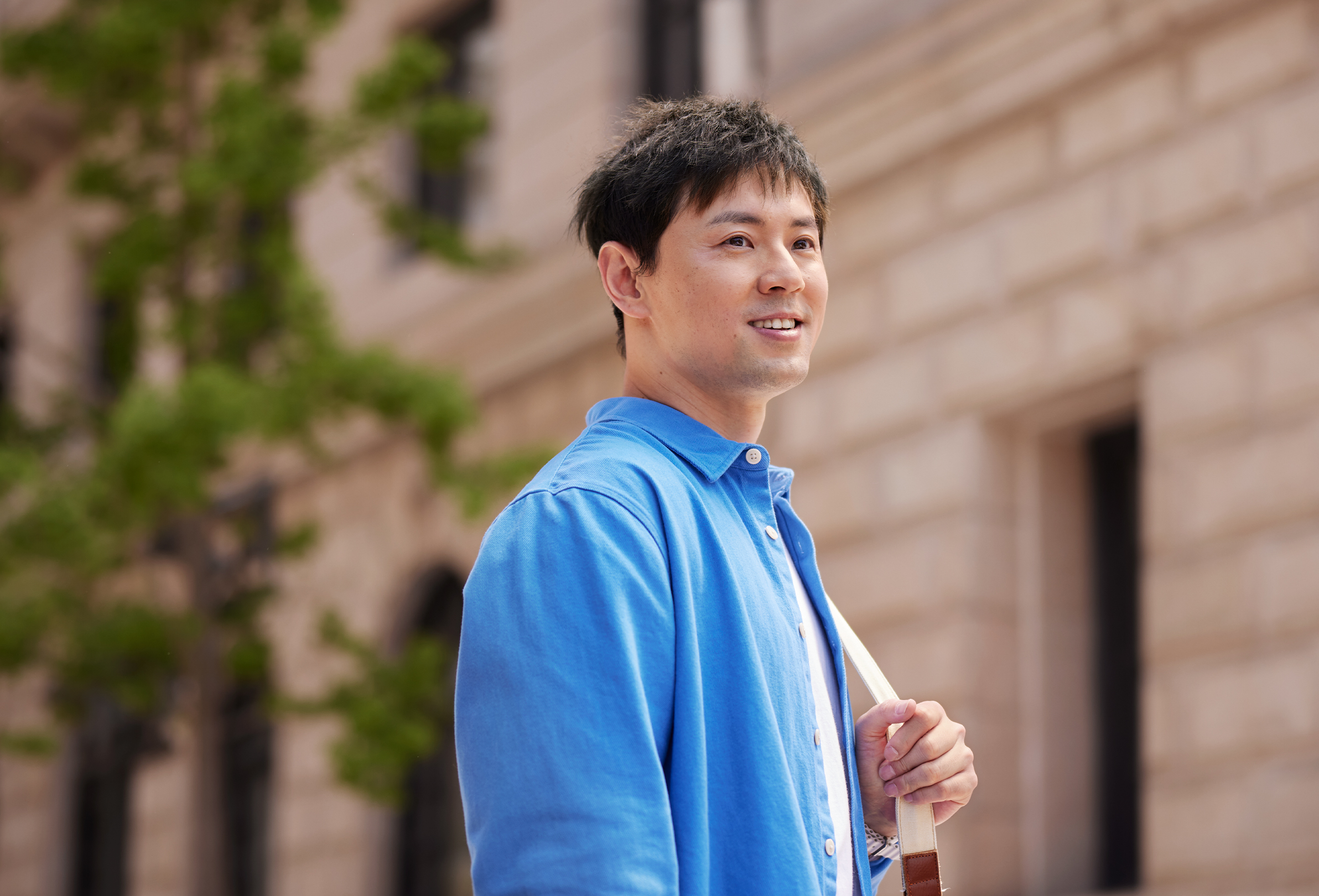 An Apple employee smiling outside a brick office building, carrying a shoulder bag.