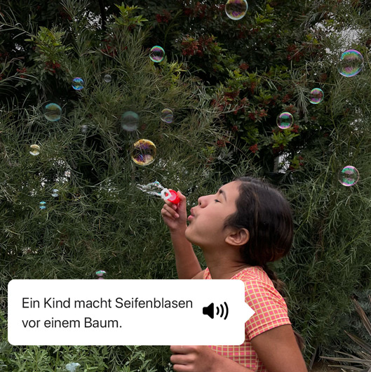 VoiceOver beschreibt ein Foto und zeigt die Sprachausgabe. „Ein Kind macht Seifenblasen vor einem Baum.“