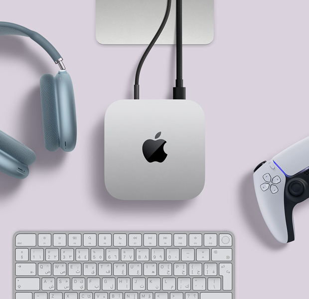 لقطة علوية لجهاز Mac mini فضي اللون، مع سلكين متصلين بالمنافذ الخلفية، وبجواره سماعات AirPods Max باللون الأزرق، ولوحة مفاتيح ماجيك باللون الفضي، ووحدة تحكم ألعاب لاسلكية