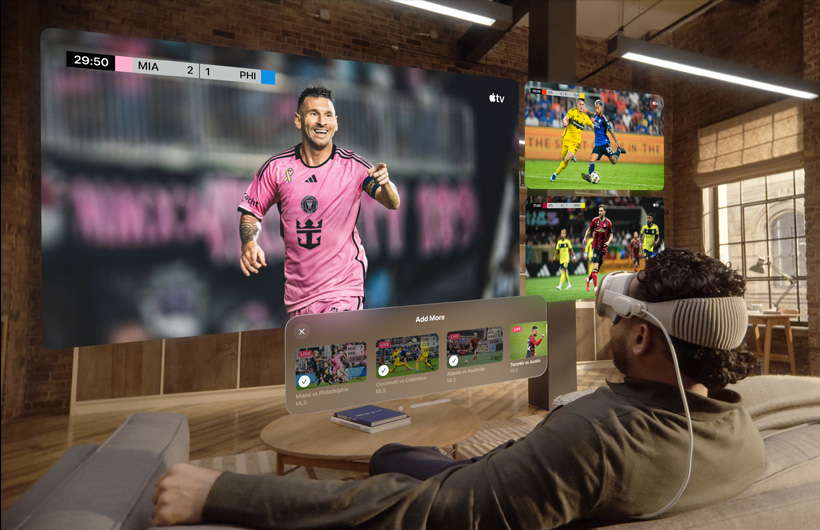 A man wearing Apple Vision Pro is sitting on a couch in a living room. Displayed in front of him are a large video screen alongside two smaller screens, each showing a different sporting event. Below the large screen is selection window that reads "Add More" along the top