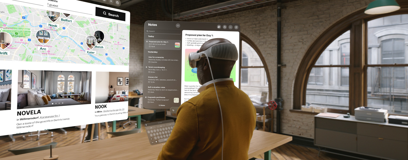 Ein Mann in leichter Seitenansicht mit einer Apple Vision Pro schaut auf mehrere Bildschirme, darunter eine Karte und die Notizen App. Er steht an seinem Schreibtisch in einem urbanen Büro.