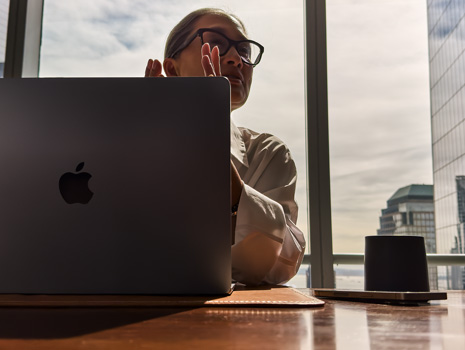 Eine Frau sitzt an einem Tisch in einem Hochhaus. Hinter ihr zeigt ein großes Fenster weitere Gebäude. Vor ihr steht ein geöffnetes MacBook.