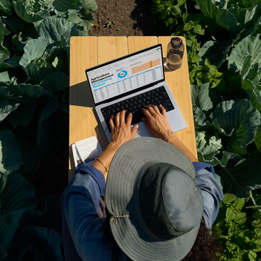 Birdseye view of a woman in a widebrim hat working on a business presentation on a MacBook, sitting at a table in a garden.