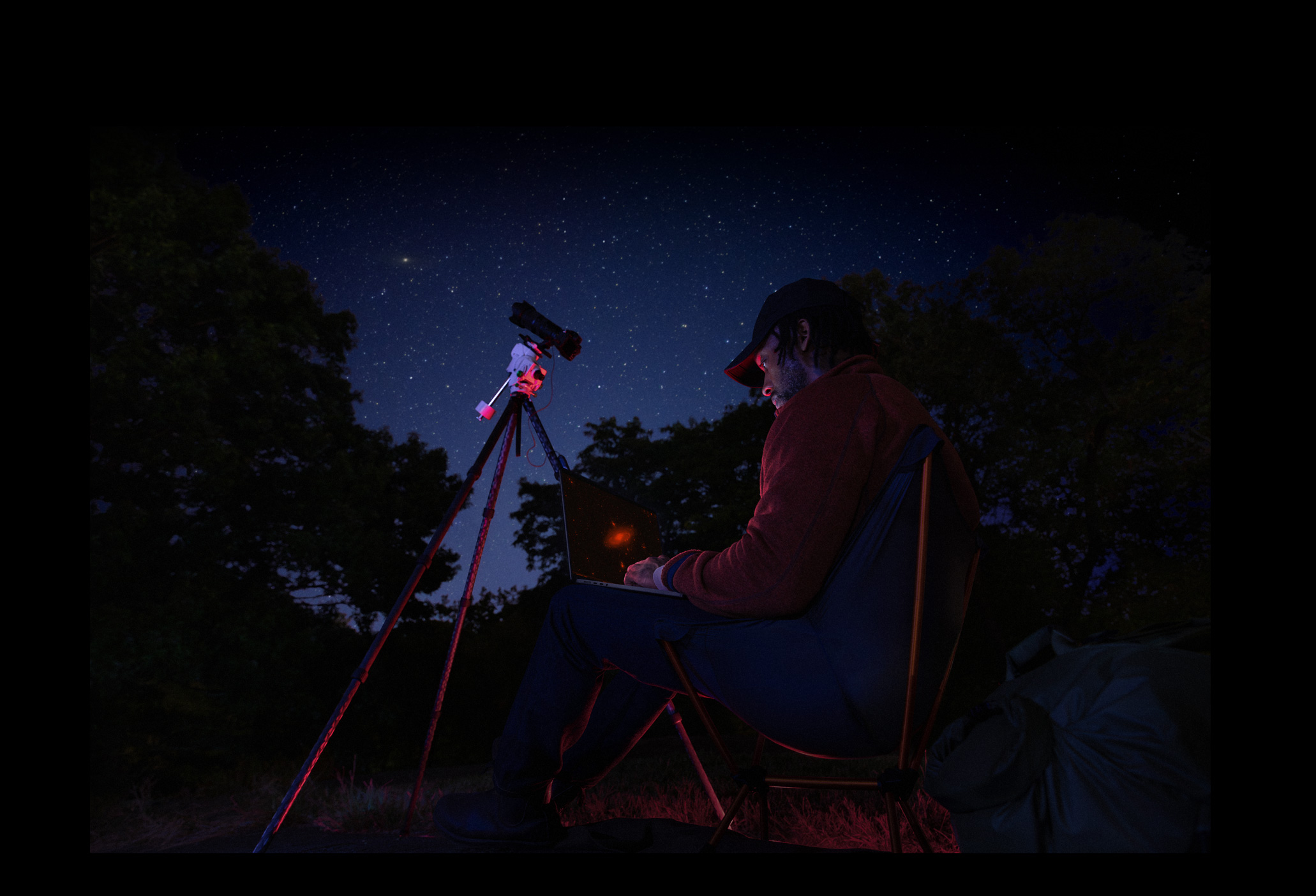 A person outside at night using a MacBook Pro on their lap, connected to a device on a tripod pointed toward the starry sky